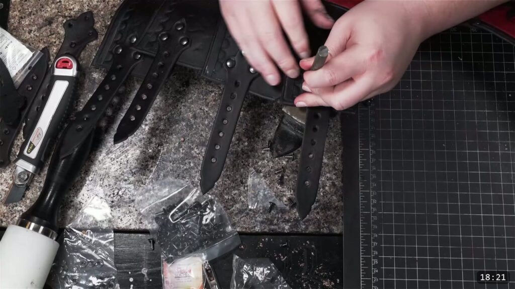 overhead of black buckle straps on a granite slab, hands setting a double cap rivet with a setter, a rotary punch and bags of rivets and hardware nearby