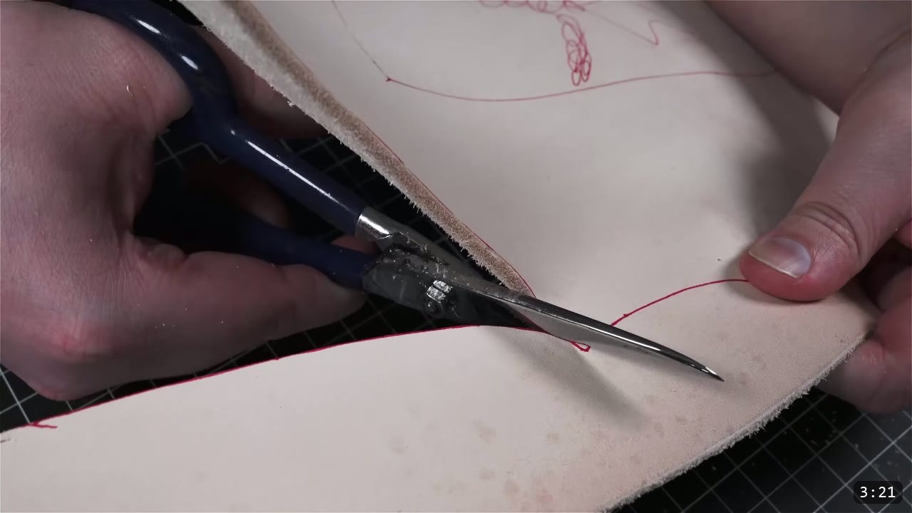 blue-handled leather shears cutting along a red traced line on a piece of tan leather on a cutting mat