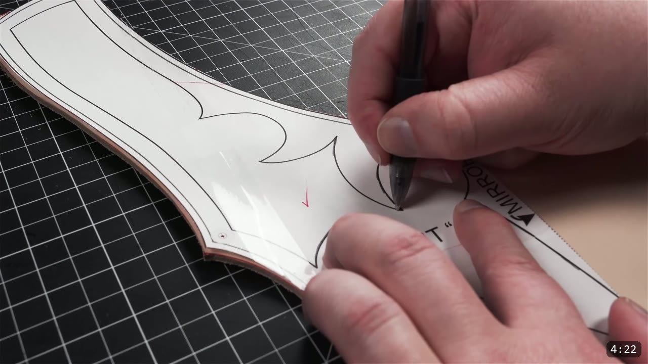 a hand tracing a barbed scalloped design element through a white paper pattern onto a cut leather piece with a black ballpoint pen
