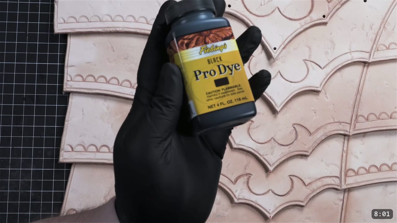 a black-gloved hand holding a bottle of Fiebing's Black Pro Dye over tan tooled breastplate plates with barbed pillow-embossed borders and punched holes