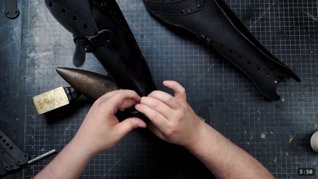 the finished pair of black leather greaves with buckle straps attached, fastening a strap over a small anvil