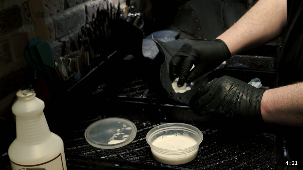 Beginner Leather Greaves Build Guide: The Easy Leg Armor to Start With 9 black-gloved hands applying a light coat of finish to the black greave with a sponge over a drying rack, a tub of finish and a spray bottle nearby