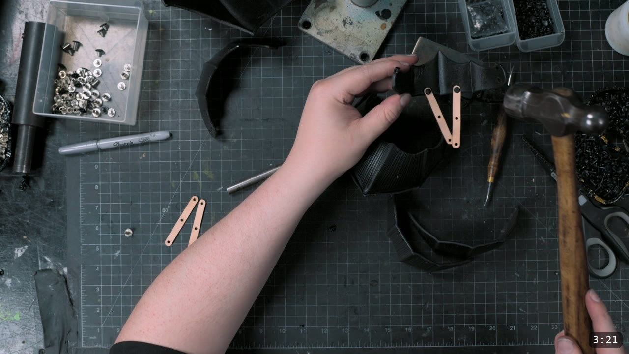 a dyed-black couter piece with pale tan retaining straps, each drilled with two holes, laid out on the mat beside a hammer
