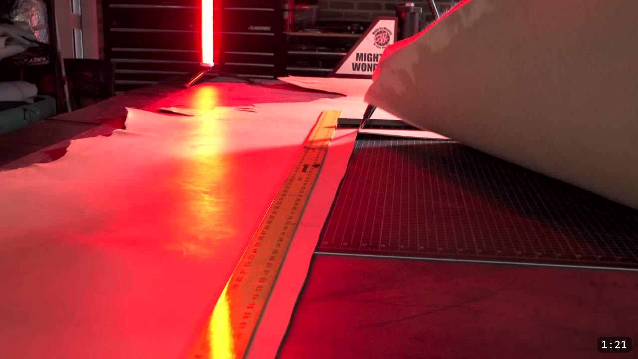 a full tan vegetable-tan hide laid flat on a cutting mat with a long steel ruler across it under red shop lighting