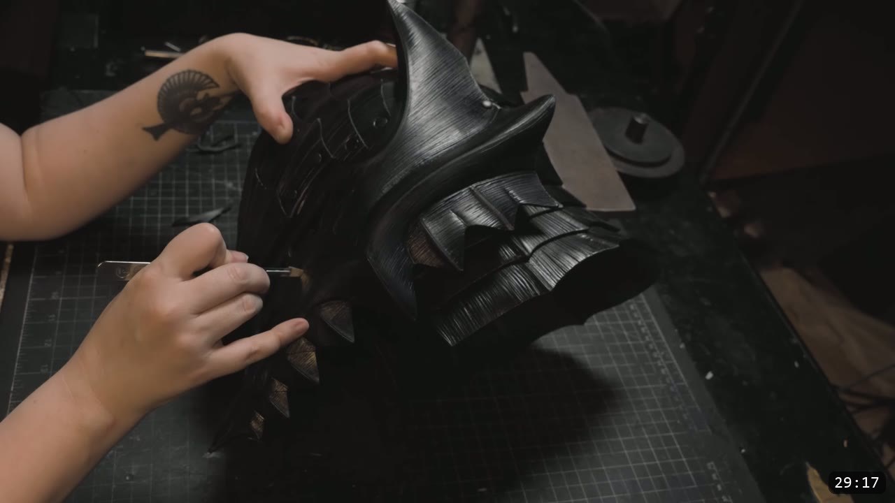 a hand refining the dyed-black faceplate with its swept horn and layered fluted edges using a fine hand tool