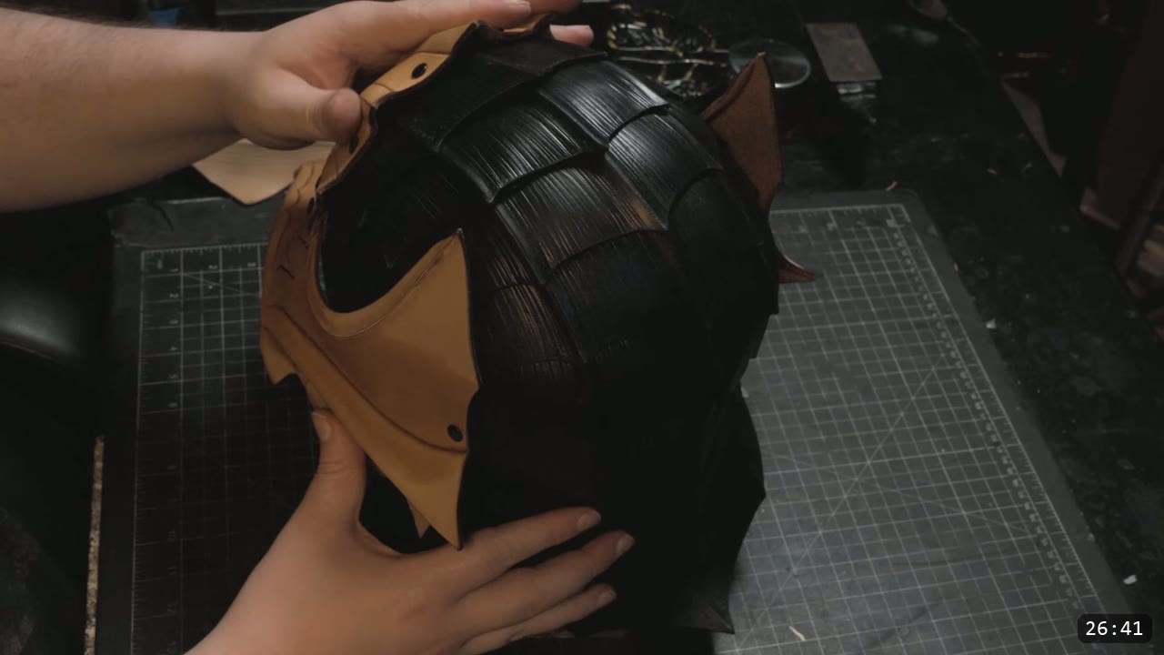 the assembled hound helmet held in two hands, a dyed-black fluted crown with a natural-tan faceplate section attached at the front