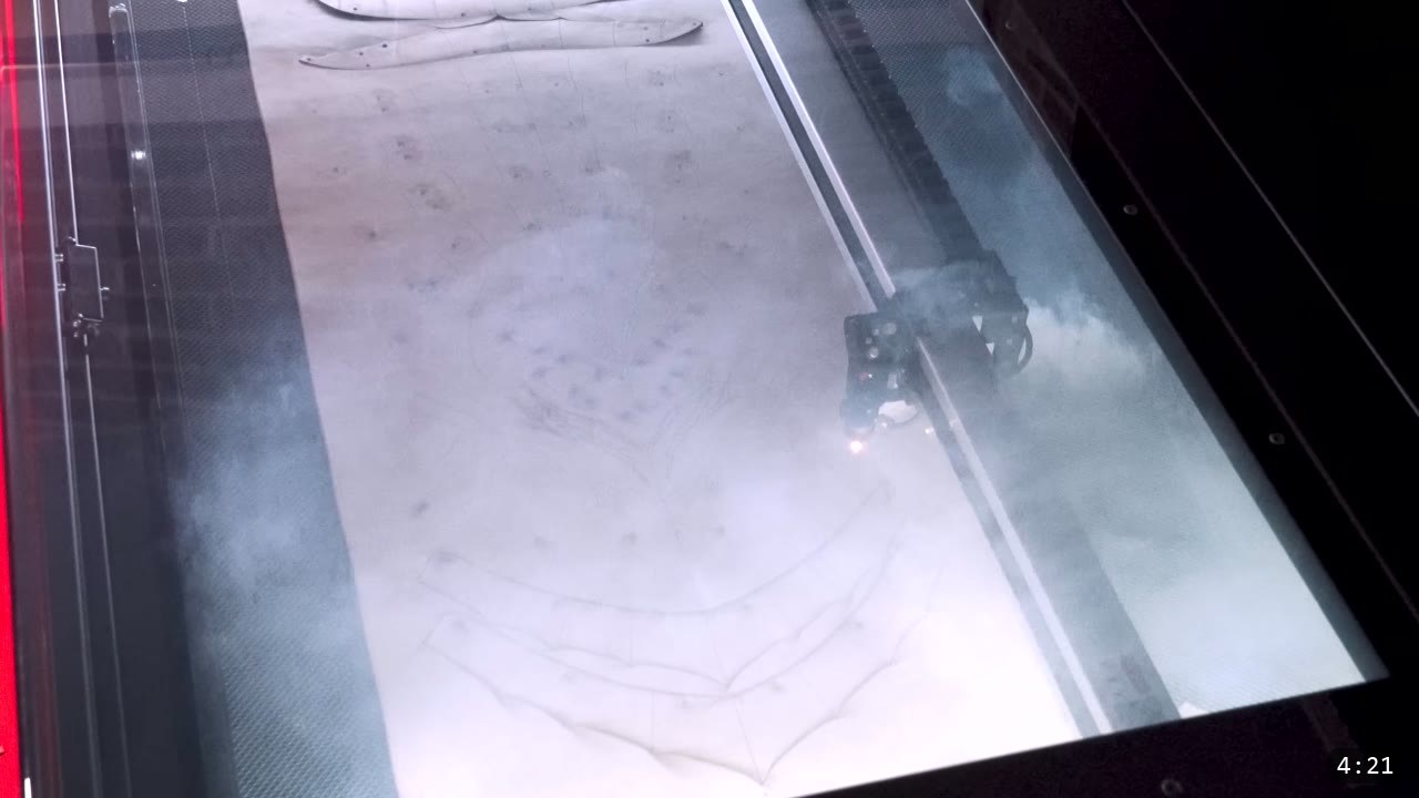 view through a laser cutter lid of the cutting head tracing helmet plate outlines with smoke rising from the bed