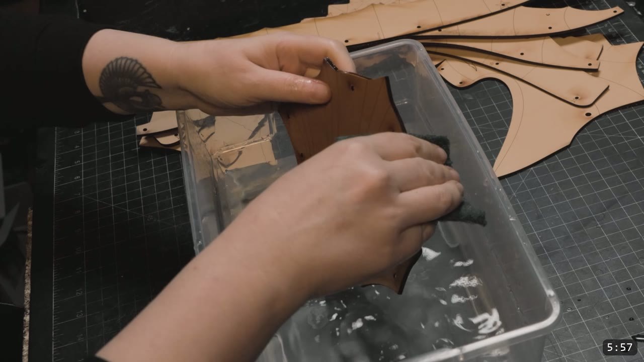 a hand scrubbing a wet-darkened leather plate with a scotch-brite pad over a tub of water, freshly cut tan plates waiting on the mat