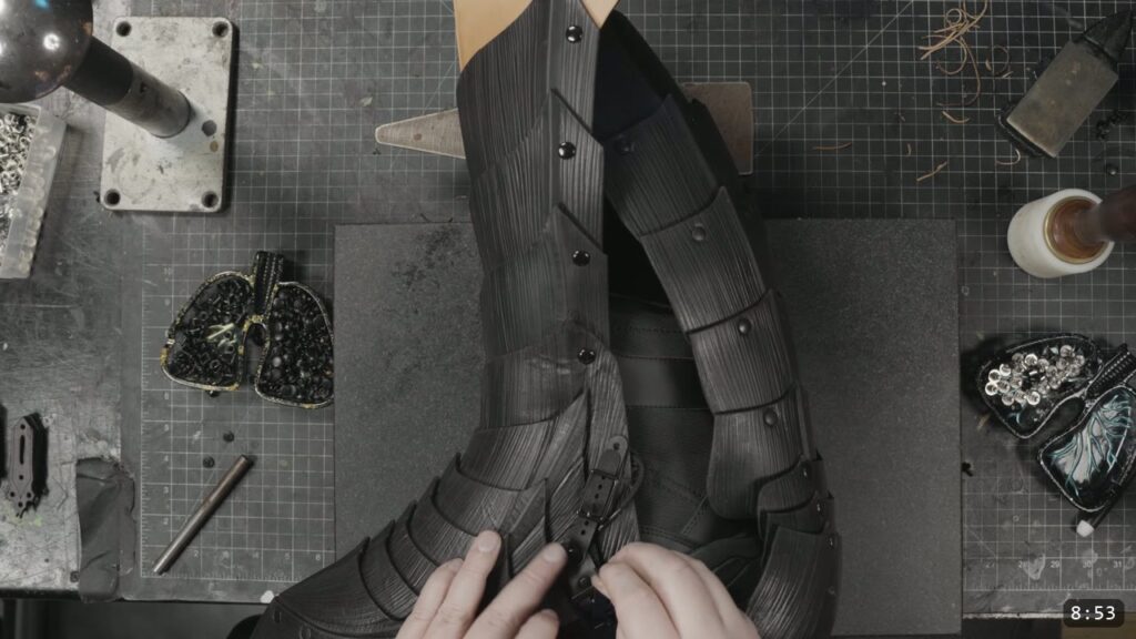 hands fastening an ankle buckle strap on the assembled black greave as it sits over a boot for a test fit