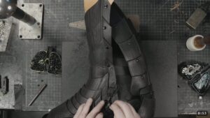 Roadmap Updates & Pre-Order Status 2 hands fastening an ankle buckle strap on the assembled black greave as it sits over a boot for a test fit
