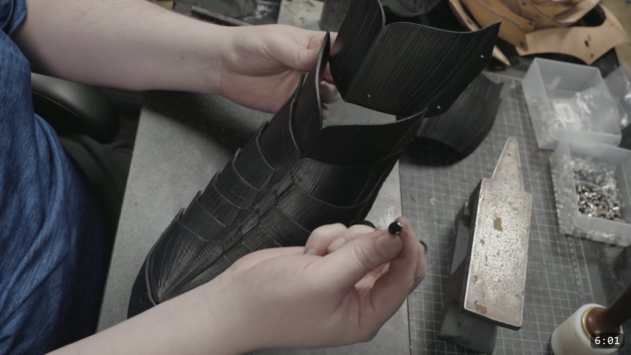 Berserk Greaves Build Guide: Leather Berserker Shin and Calf Armor 10 hands setting a black double-cap rivet on the black fluted front greave, a forming stake and hardware bins nearby