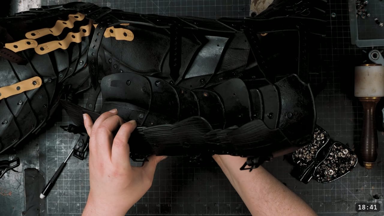 Berserk Leg Armor Build Guide: Articulated Leather Cuisses, Knees, and Greaves 12 The assembled black leg armor laid flat, natural retaining straps threaded through the inside of the overlapping plates, hands adjusting a lower fluted section