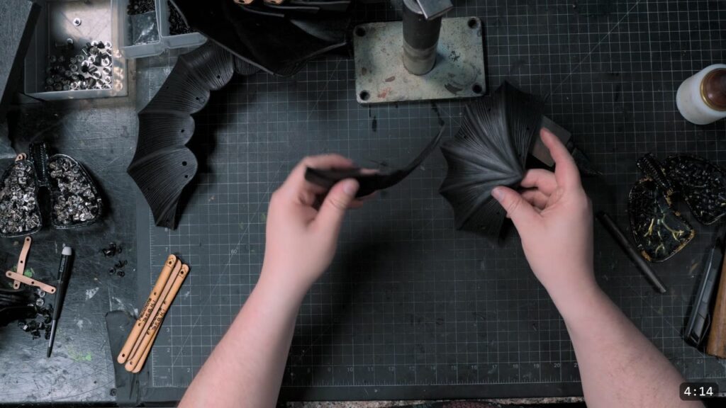 overhead view of dyed-black fluted pauldron plates being handled, with a tray of rivets and a wing divider on the bench