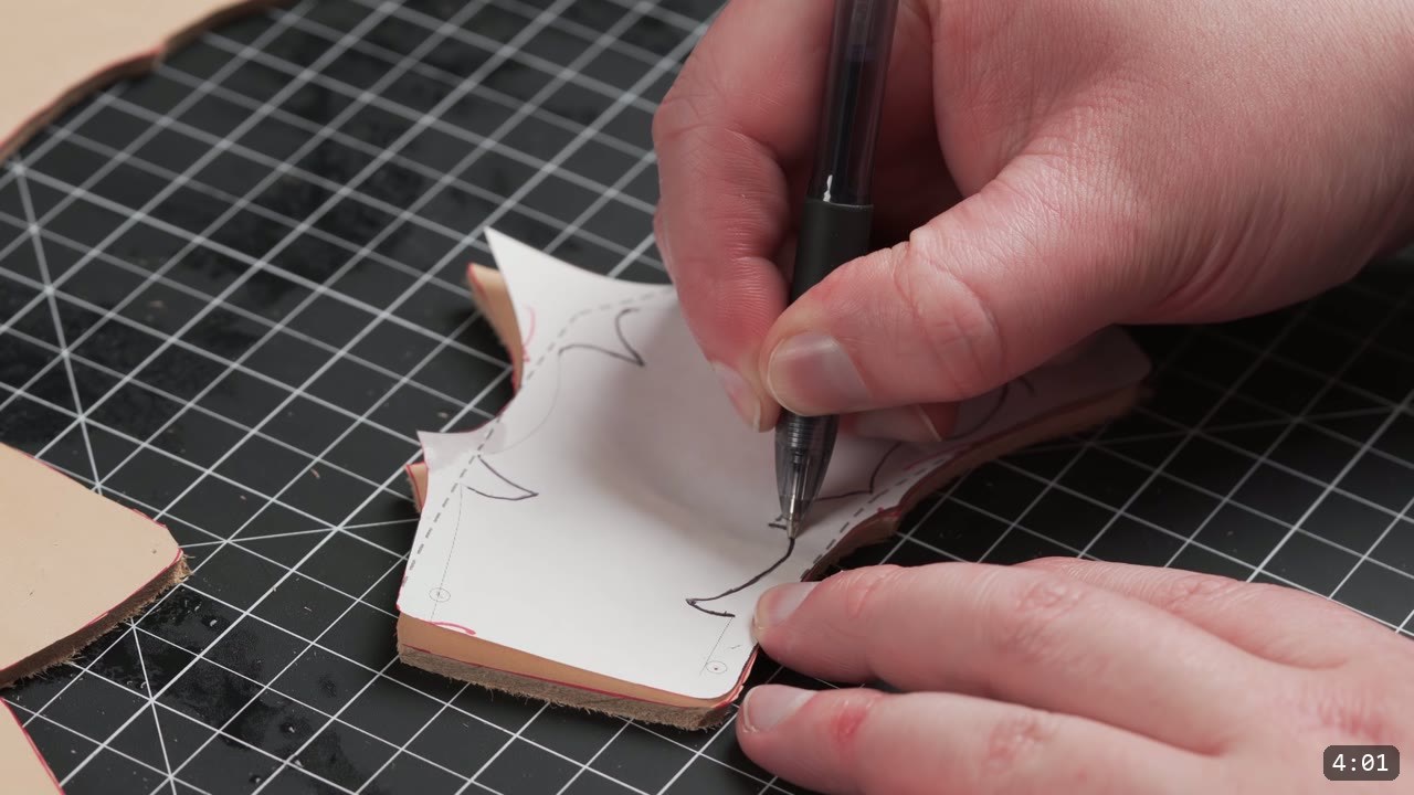 laying a paper pattern over a cut leather piece and tracing the design lines with a ballpoint pen on a cutting mat
