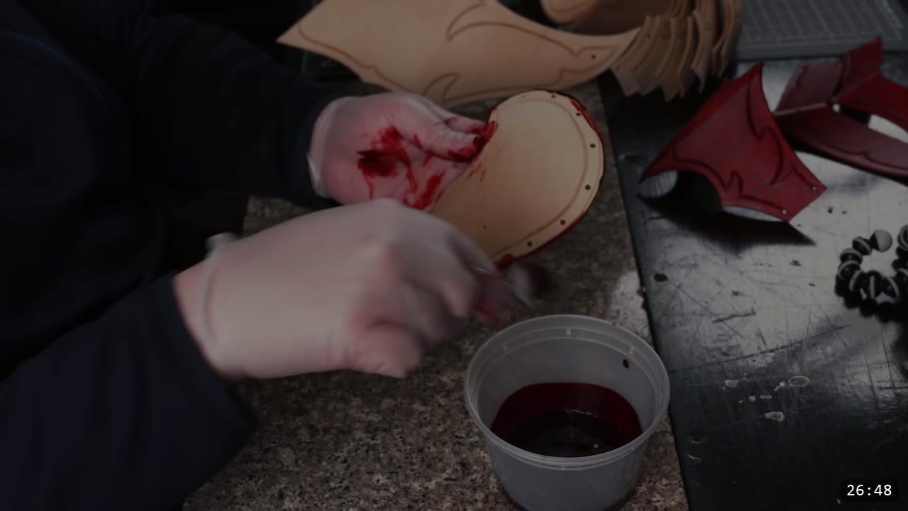 gloved hands working red water stain into a punched leather cheek piece beside a tub of red dye, finished red pieces drying to the side