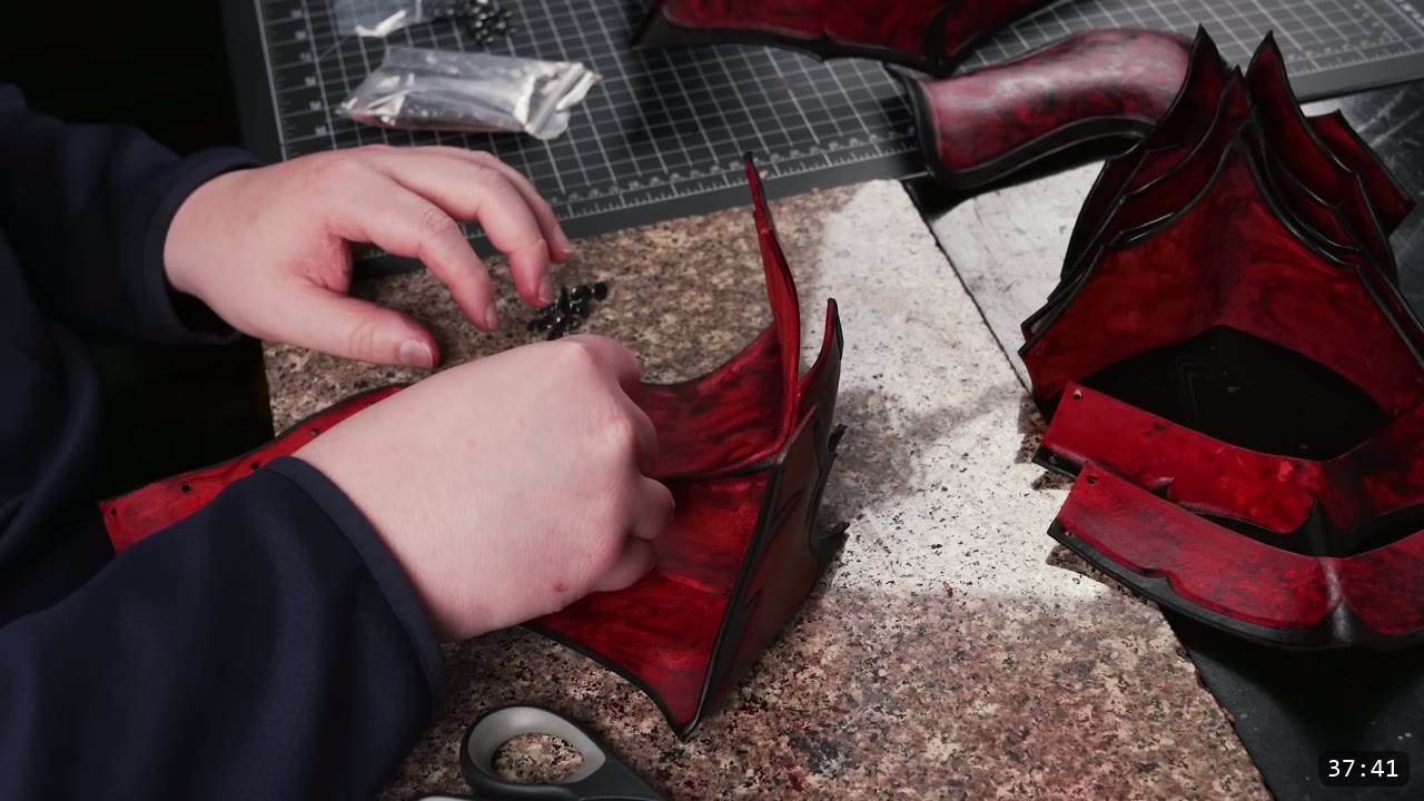 two hands riveting the marbled red-and-black helmet core together with gunmetal rivets, a bag of rivets and shears nearby