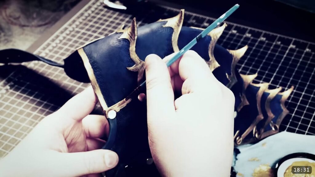 a small brush applying metallic gold to the raised barb edges of the dark-blue spaulder, several barbs already highlighted gold