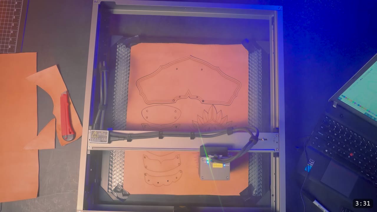 a large sheet of tan vegetable-tan leather on the laser bed with the cuff plate and feather-crest pieces cut and outlined, laptop running the cut file to the right