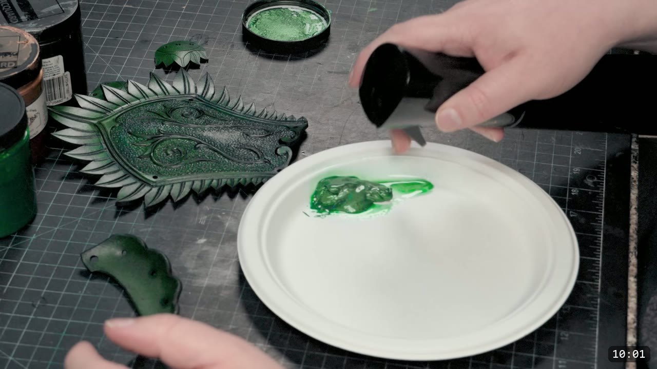a hand loading paint onto a white paper plate beside a finished metallic-green feather-crest cuff piece with a tooled swirl, paint jars to the left