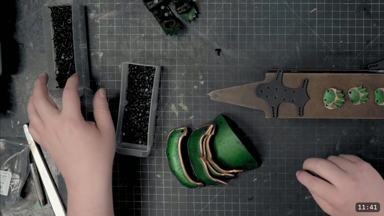top-down view of two organizer boxes of black double-cap rivets, a riveted knuckle band, and green finger scales being fitted