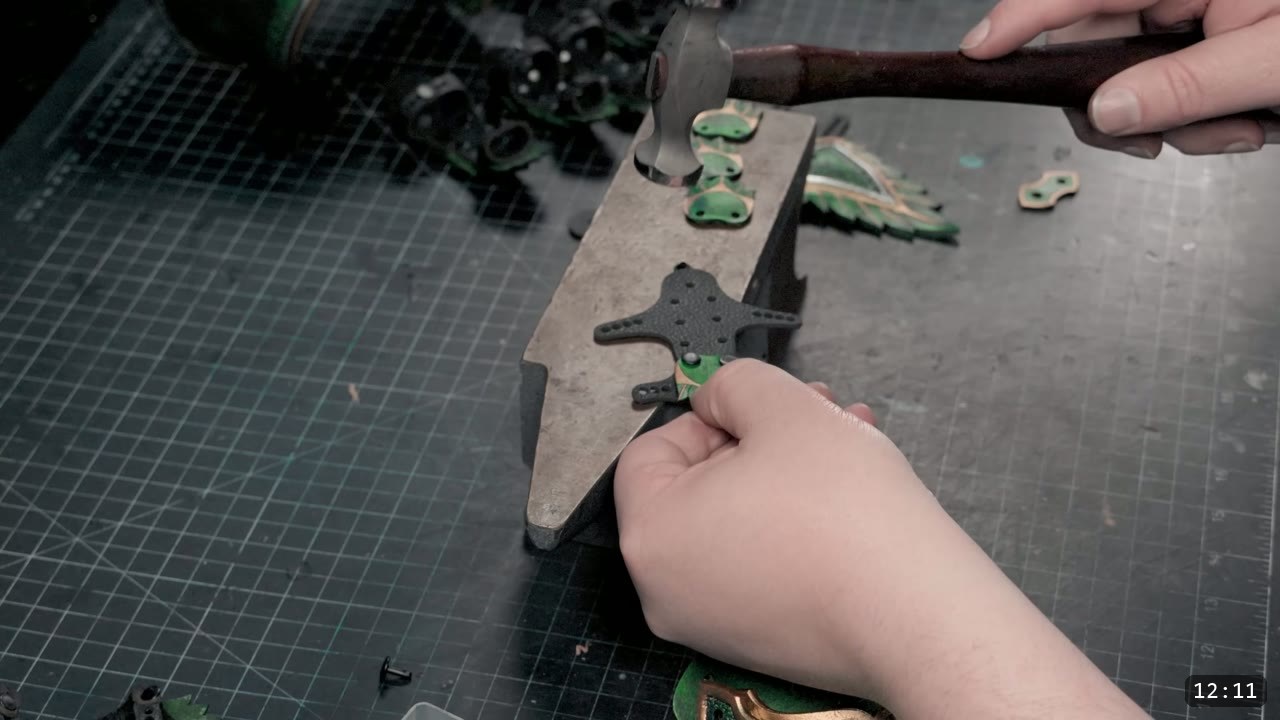 a small hammer setting a rivet on a green-and-black finger scale held against a small steel horn anvil, more assembled finger scales behind