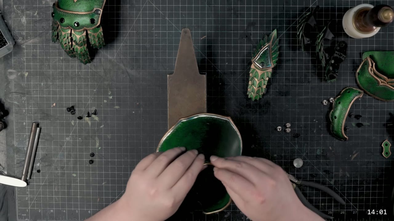 hands bending a dished metallic-green cuff piece near a steel bench stake, a completed green scaled finger unit and thumb piece on the mat