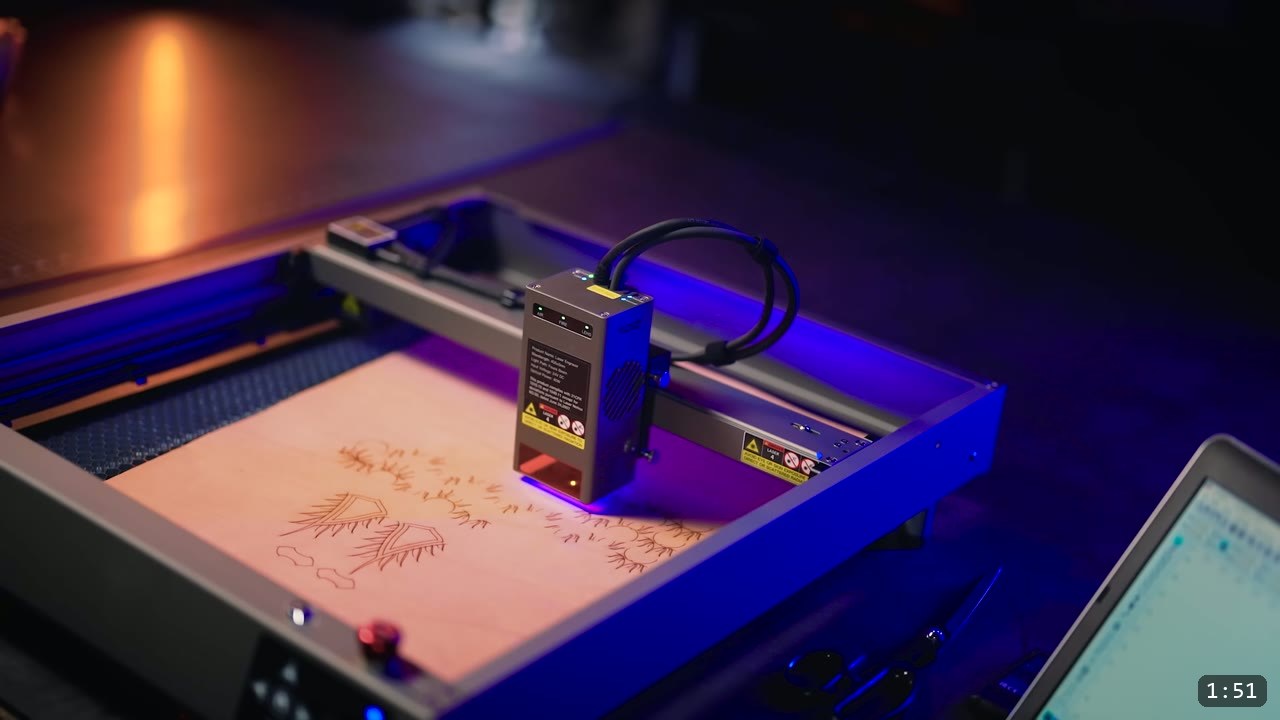 a desktop laser head on its gantry engraving a test pattern of feather motifs, honeycomb bed below and a laptop running the cut software