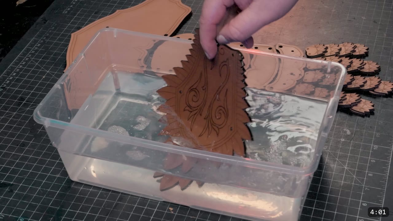 a hand dunking a laser-cut feather piece with an engraved swirl into a clear plastic bin of water, other cut pieces resting on the mat