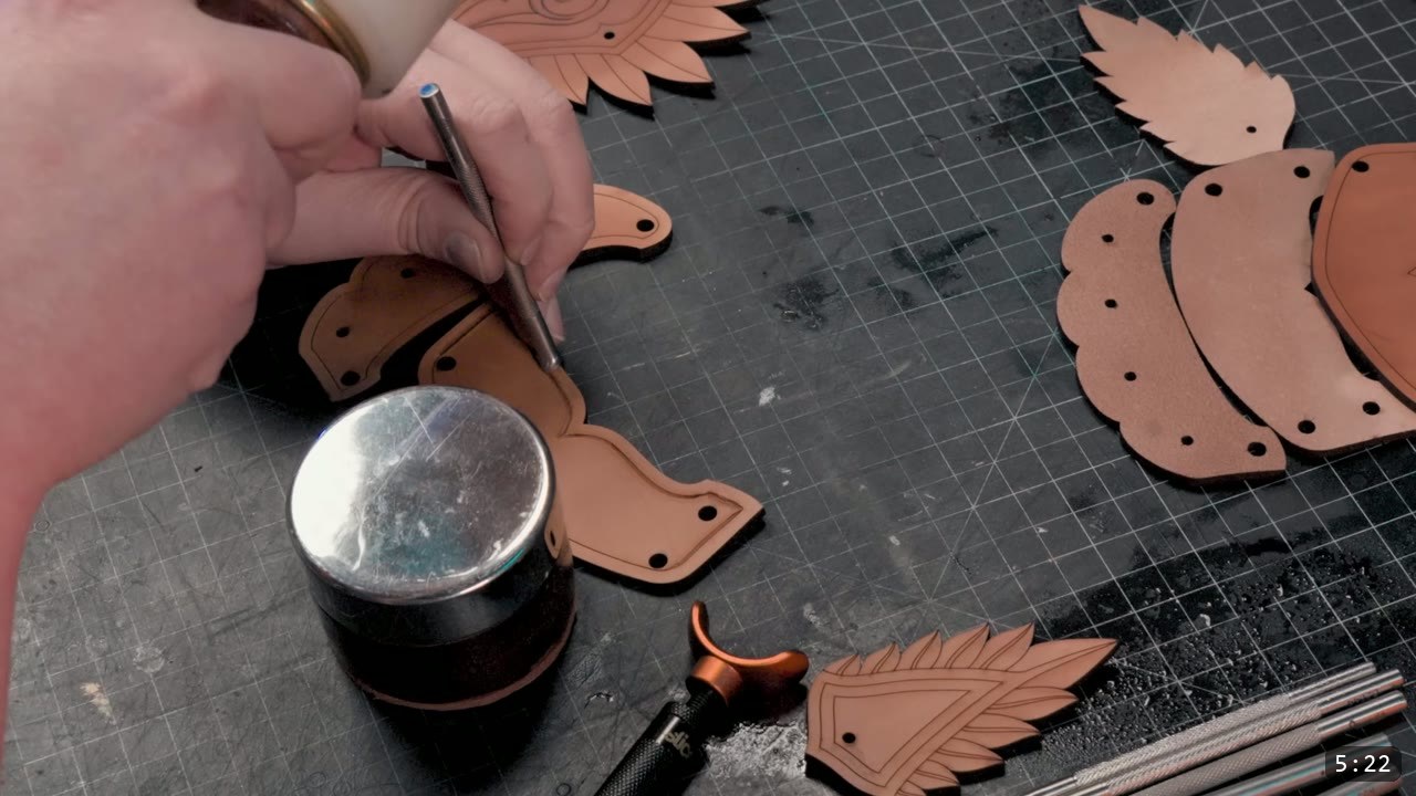 a hand working a carved cuff piece with a modeling tool on a cutting mat, a round steel anvil and cut cuff bands nearby