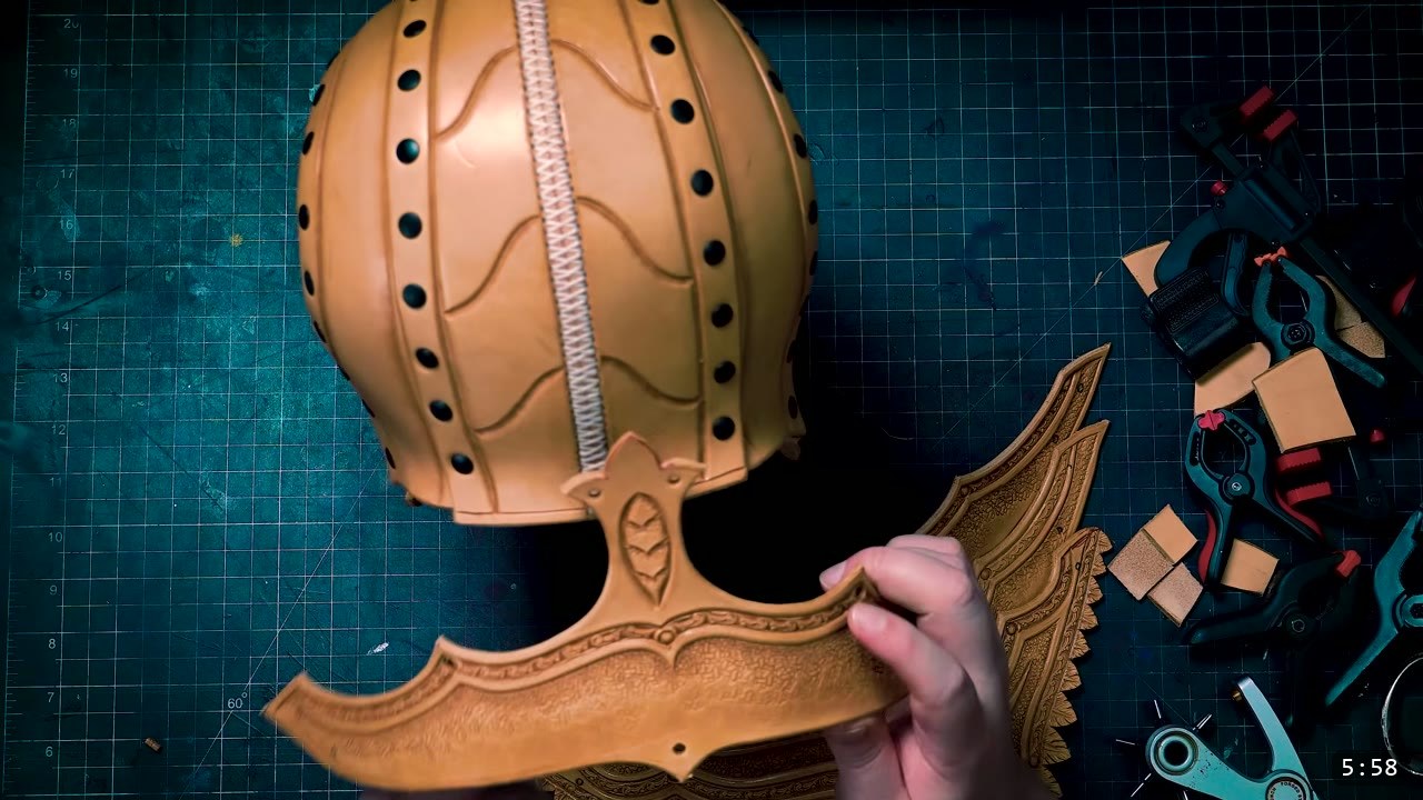 Leather Elven Helmet Build Guide: Visor, Comb, Tail, and a Green Metallic Finish 2 the assembled tan Elven Lord helmet core with a laced center seam, held above a tooled tail band and a carved feathered side plate on a cutting mat