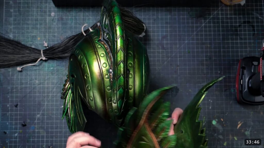 the near-finished metallic green Elven Lord helmet with a feathered comb and a black horsehair plume tied at the back, hands fitting a green feathered side plate