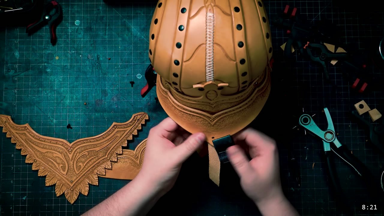 Leather Elven Helmet Build Guide: Visor, Comb, Tail, and a Green Metallic Finish 6 hands attaching a dark strap to the base of the laced helmet core over a tooled back band, a carved feathered plate resting to the left and rotary punch pliers to the right