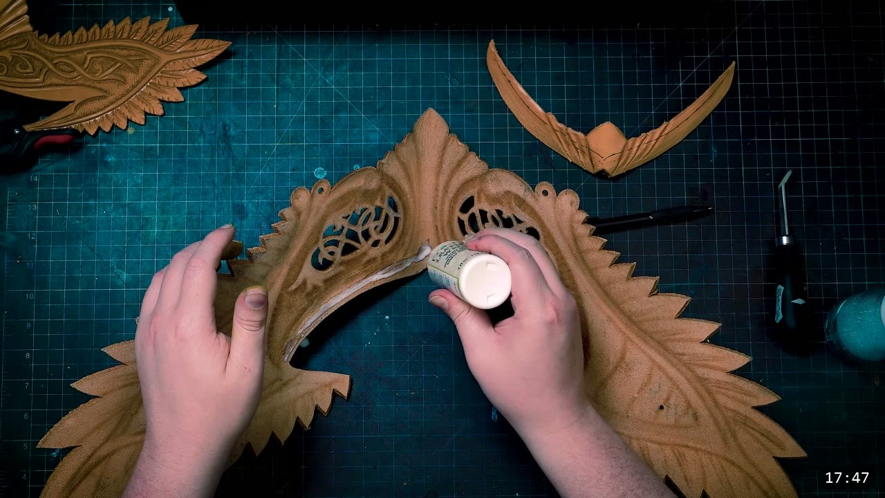 Leather Elven Helmet Build Guide: Visor, Comb, Tail, and a Green Metallic Finish 9 hands applying white leather glue along the inside edge of a carved filigree visor plate with openwork scrollwork, other feathered pieces and an awl nearby