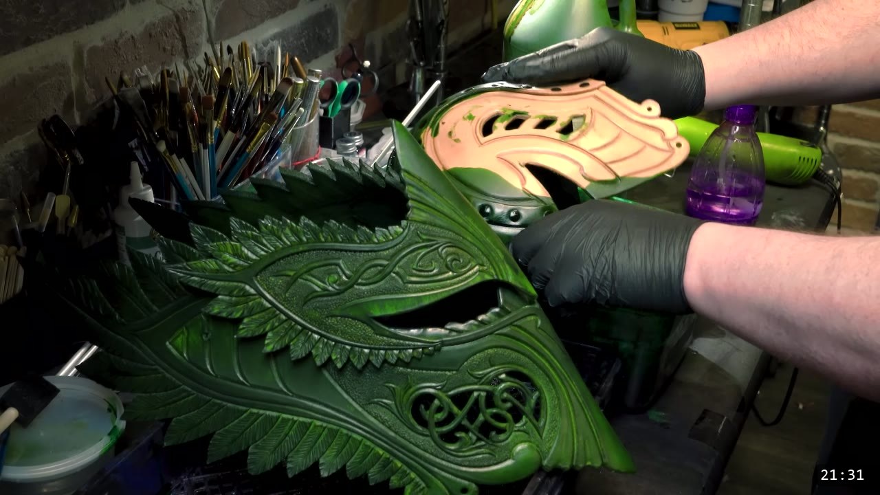Leather Elven Helmet Build Guide: Visor, Comb, Tail, and a Green Metallic Finish 10 gloved hands immersion dyeing a carved feathered plate deep green, a green-dyed faceplate and a spray bottle on the bench, brushes in jars behind