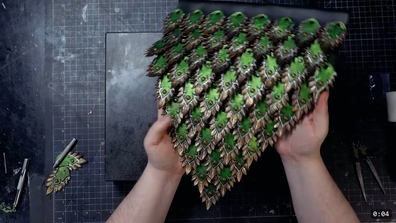 finished elven leather scale panel held in both hands, green scales with bronze feathered edges and a stud in each center, arranged in a triangular skirt over a cutting mat
