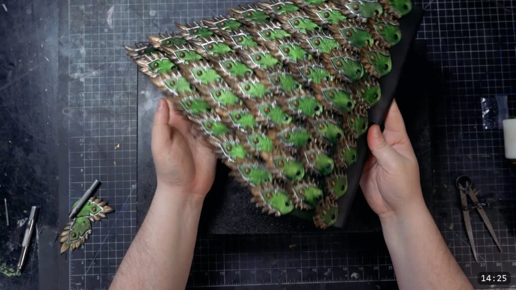 both hands holding the finished triangular scale panel, overlapping rows of green scales with silver and bronze edges and copper rivets