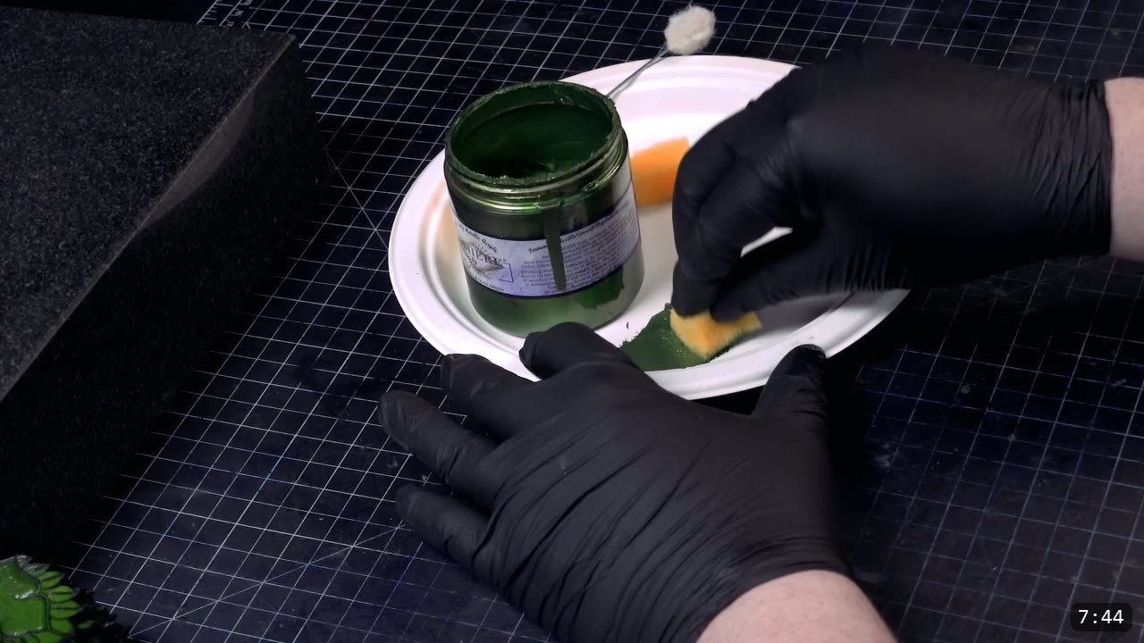 gloved hands applying green color to a scale with a small sponge on a paper plate, an open jar of green beside a wool dauber