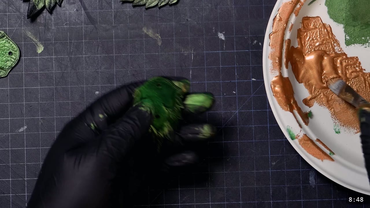 gloved hand holding a green-painted scale beside a plate loaded with metallic bronze and green paint, a brush picking up bronze