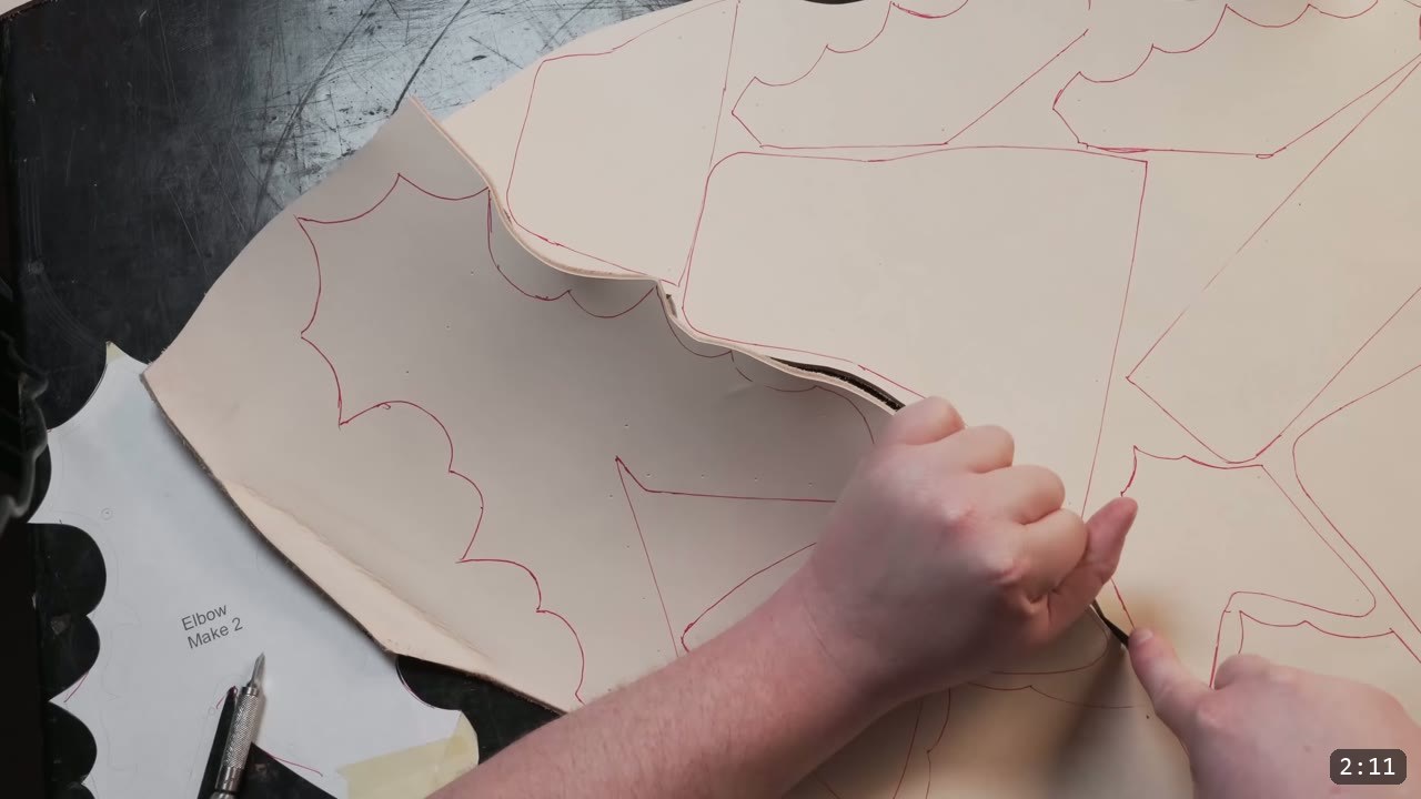 Leather Arm Armor Build Guide: Fantasy Vambrace, Rerebrace, and Elbow 4 a tan veg-tan sheet with fantasy arm-armor pieces traced in red, separating the cut parts by hand with a razor and a paper template nearby