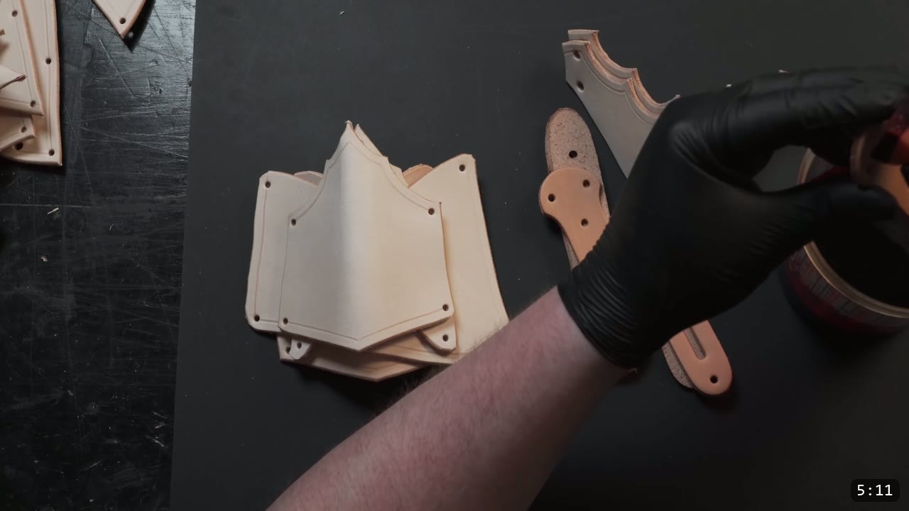 Leather Arm Armor Build Guide: Fantasy Vambrace, Rerebrace, and Elbow 7 undyed tan arm-armor pieces and straps laid out on a black bench, a gloved hand beginning the dye step