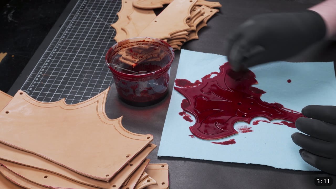 Leather Cuisses and Knees Build Guide: Articulated Upper-Leg Armor 7 a gloved hand brushing red oil dye onto a bat-wing knee fan piece over blue paper, a cup of red dye and undyed tan pieces beside it