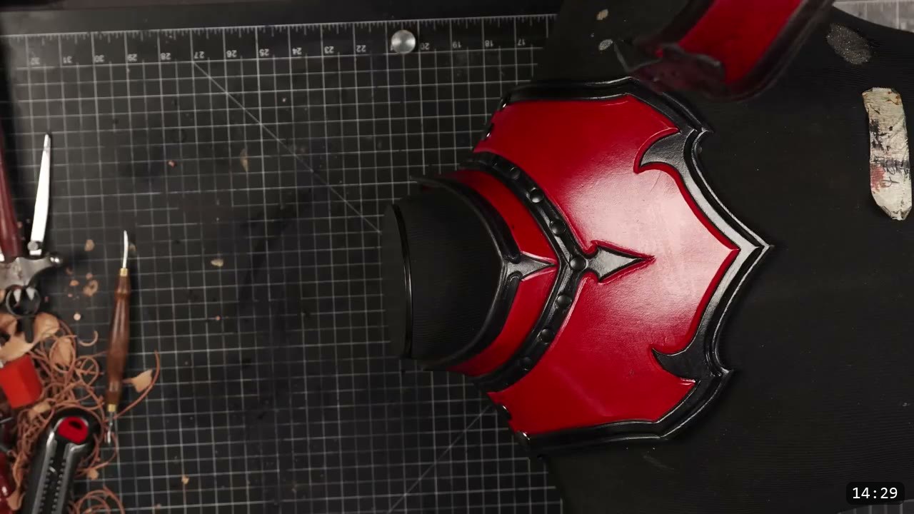 Leather Gorget Build Guide: A Fantasy Armor Neck Guard, Step by Step 2 finished red leather gorget front plate with black tooled borders and a carved center motif, resting on a cutting mat beside a swivel knife and box cutter