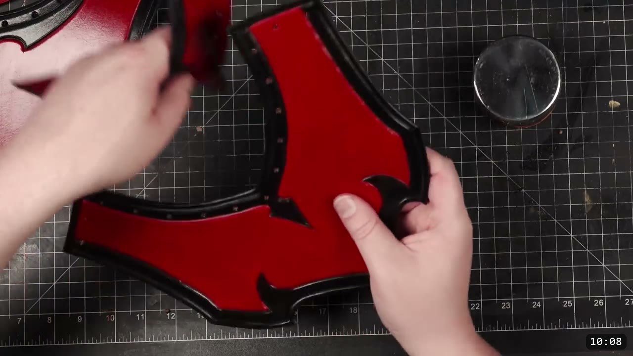 Leather Gorget Build Guide: A Fantasy Armor Neck Guard, Step by Step 11 hands holding the dyed red gorget front plate flat, its black border set with a row of rivets, a round glass weight to the side