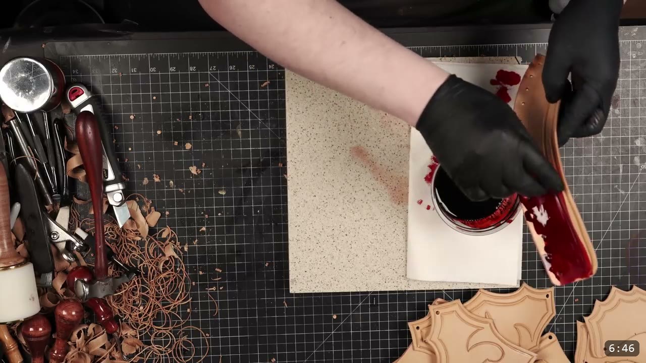 Leather Gorget Build Guide: A Fantasy Armor Neck Guard, Step by Step 9 black-gloved hands staining a cut tan gorget strap red in a jar of dye, leather scrap curls and stamping tools on the left