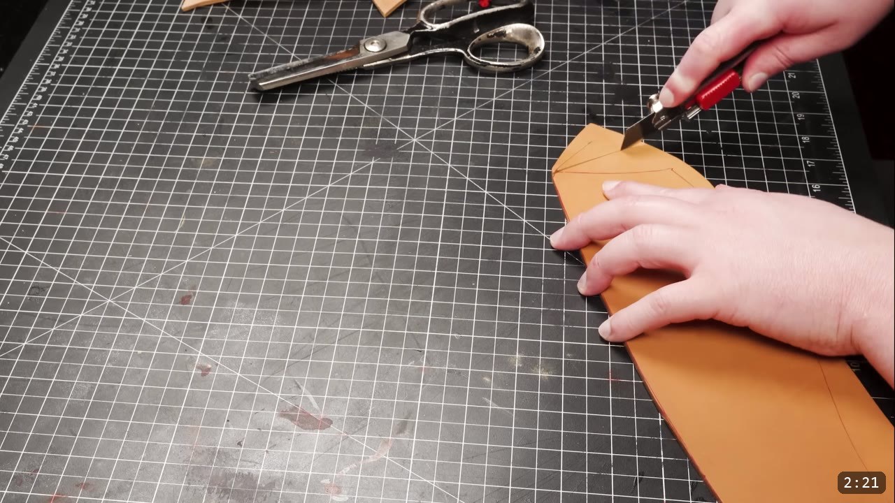 Leather Greaves Build Guide: From Flat Pattern to Antiqued Leg Armor 5 cutting a long curved edge of a tan leather greave panel with a red-handled box cutter, heavy shears resting on the cutting mat