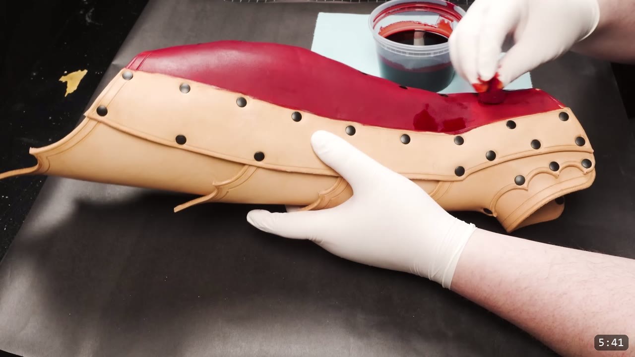 Leather Greaves Build Guide: From Flat Pattern to Antiqued Leg Armor 10 brushing a solid red oil dye across the top of the fully riveted tan greave, a cup of red dye alongside on black paper