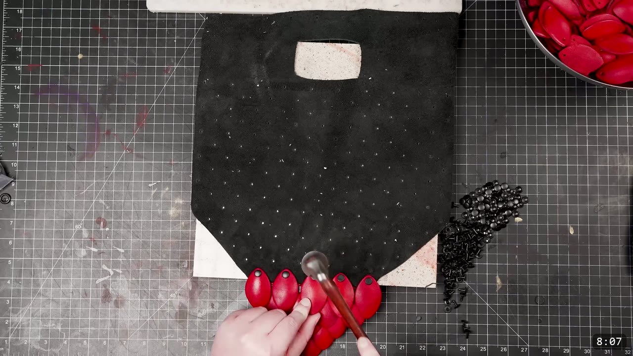 Leather Scale Armor Build Guide: A Flexible Scale Skirt from Scrap Leather 9 the cut black backing panel with punched holes and buckle slot, first red scales being riveted at the bottom edge next to a pile of black rivets and a bowl of red scales