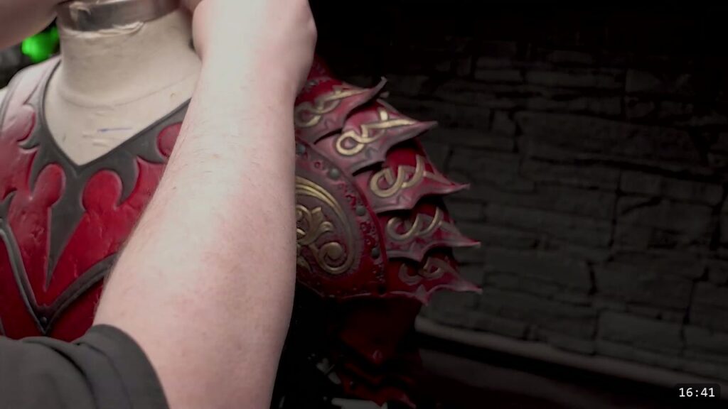 a finished deep-red spaulder with barbed overlapping plates and gold-highlighted scrollwork, mounted at the shoulder of a mannequin wearing the matching red fantasy breastplate against a stone wall