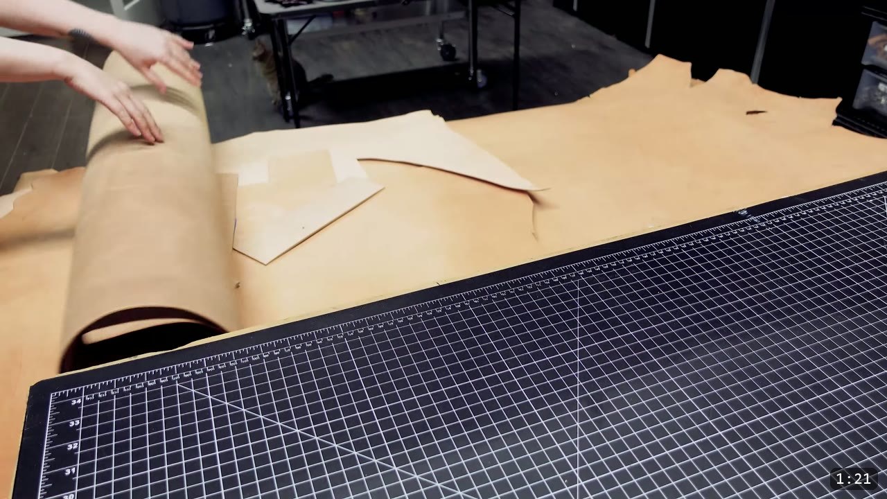 rolling out a full vegetable-tan side on a large gridded cutting mat with an arrow-marked paper pattern piece laid on the leather