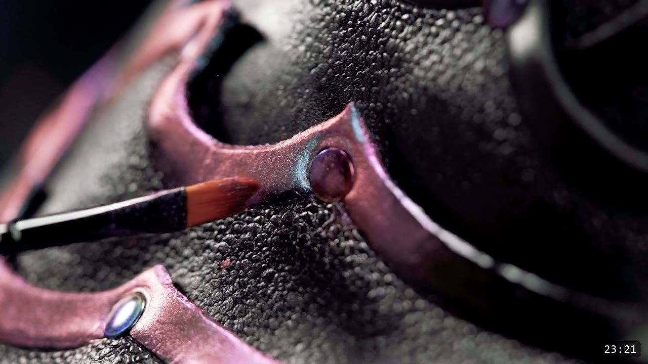brushing color-shift pigment onto the raised tooled border of the dyed-black corset, a set rivet catching pink and teal light